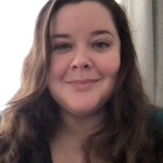 head shot of woman with long, brown wavy hair, black v-necked sweater, in front of window