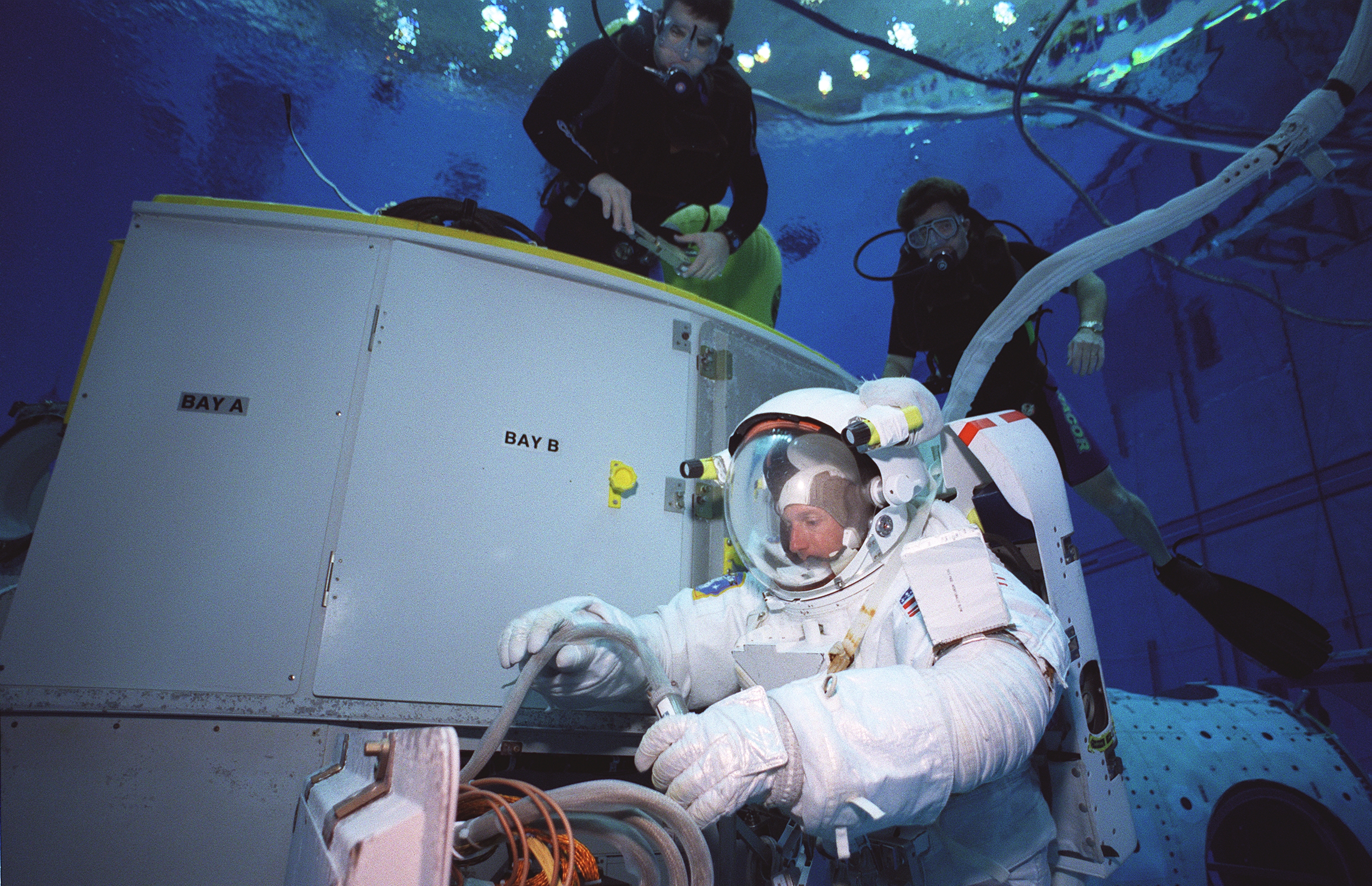 Clad in a spacesuit, an astroanut in an underwater tank pulls a tube from a replica of Hubble's bay doors. The labels "Bay A" and "Bay B" are visible on the doors. Two black-suited divers hover above him in the tank.