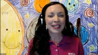 Video still frame of a zoom call focused on a woman with long dark hair, smiling, and sitting in front of a child's colorful crayon drawing of the solar system.