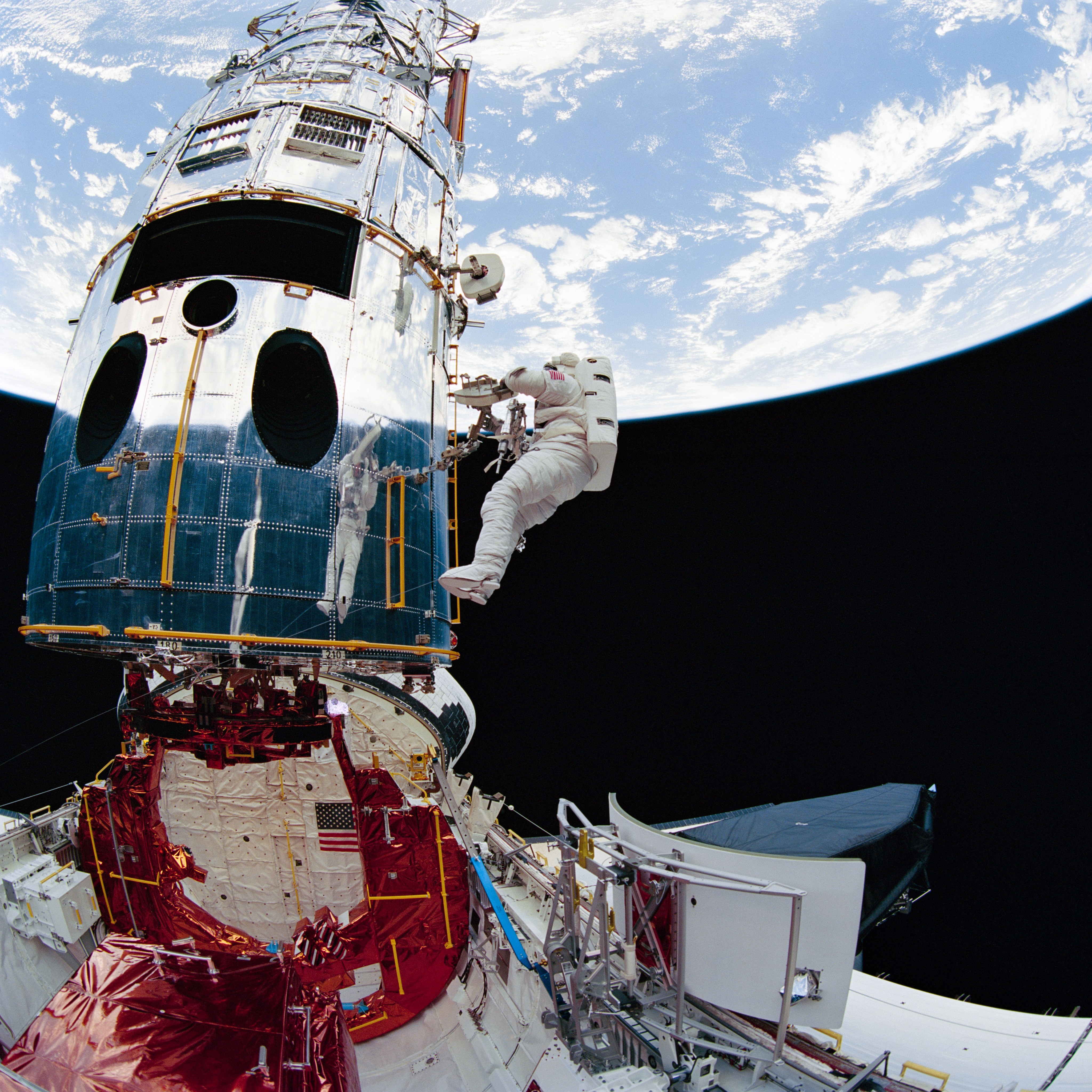 With Earth in the background, an astronaut works on Hubble and is reflected in the shiny surface of the telescope. Below him, the space shuttle's metallic, red-colored Flight Support System helps hold the telescope in position, and the newly removed Wide Field Planetary Camera 1 is stowed temporarily nearby.