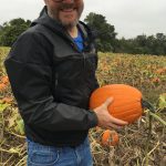 bearded man with glasses in black ball cap, windbreaker, jeans, holding a pumpkin in a pumpkin patch