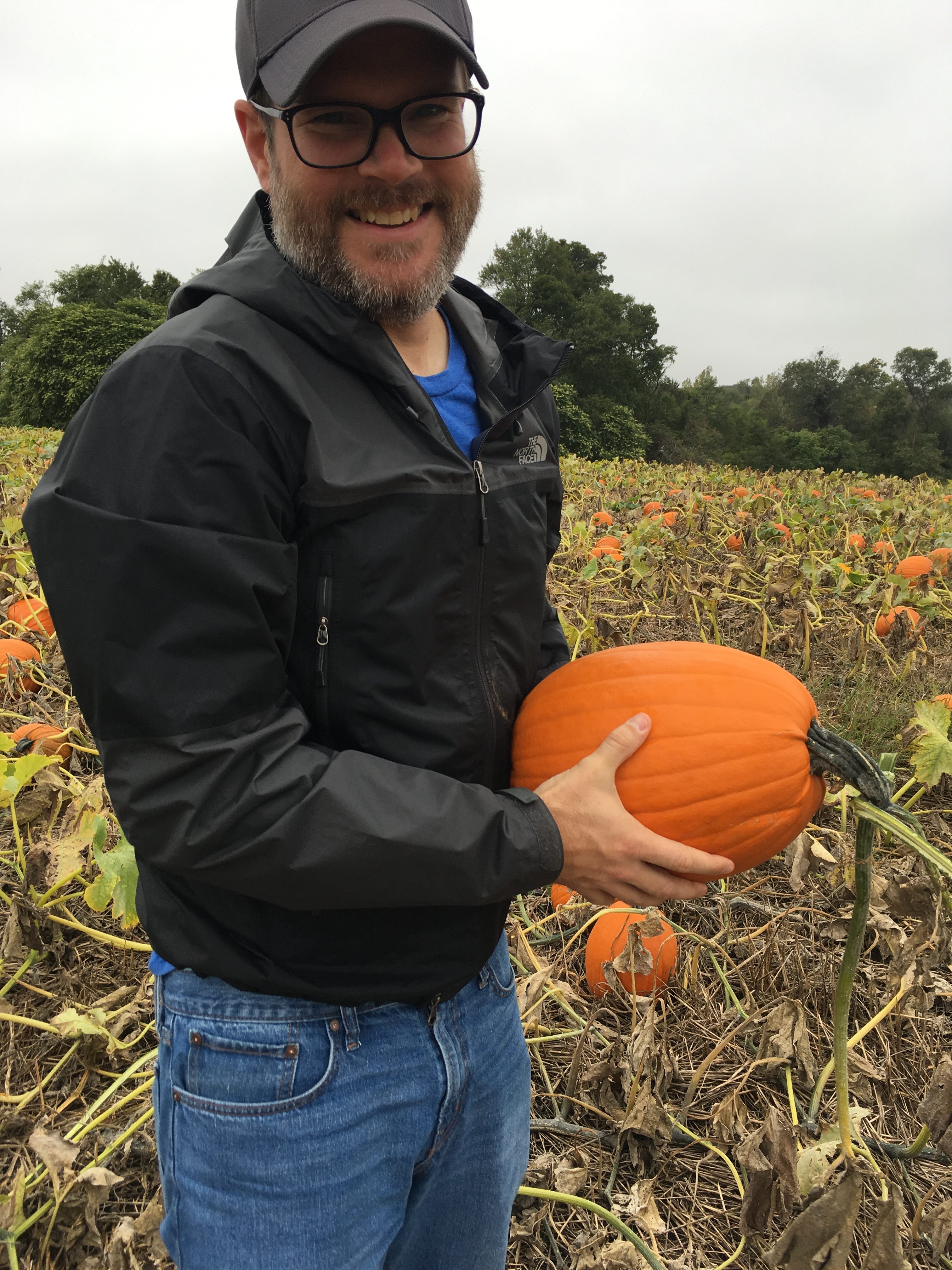 bearded man with glasses in black ball cap, windbreaker, jeans, holding a pumpkin in a pumpkin patch