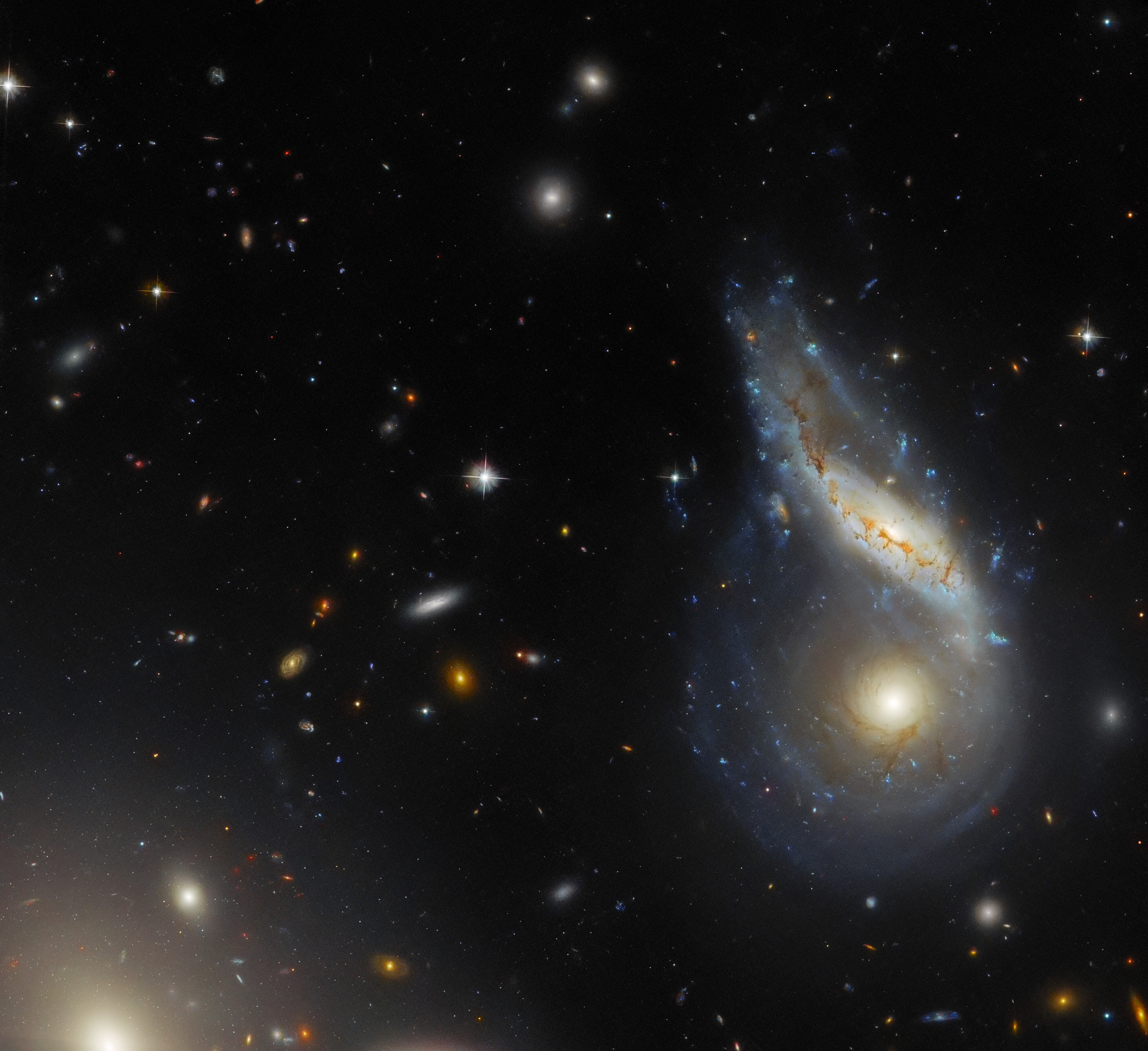 Two spiral galaxies are merging together at the right side of the image. One is seen face-on and is circular in shape. The other seems to lie in front of the first one. This galaxy is seen as a disc tilted away from the viewer and it is partially warped. In the lower-left corner, cut off by the frame, a large elliptical galaxy appears as light radiating from a point. Various small galaxies cover the background.