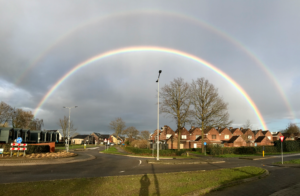 The image shows a landscape of a neighborhood with grass areas, streets, and some houses to the left at the horizon. There are som trees in the center as well as a lamp post. The sky is cloudy and a gray color. The featured part of the image isa double rainbow, spanning from the entire left to right of the image. The inner rainbow is brighter while the outer rainbow is faint.