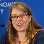 woman with layered, mid-length blonde hair, red glasses, speaking a mic with dark blue blouse, cobalt-blue pendant necklace