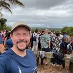 man with goatee, wearing tan ball cap and blue T-shirt, taking selfie in front of crowd watching weather system in the distance