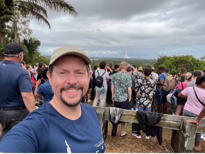 man with goatee, wearing tan ball cap and blue T-shirt, taking selfie in front of crowd watching weather system in the distance