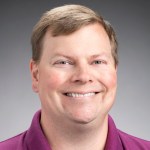 headshot of smiling blonde man in magenta-colored collard shirt