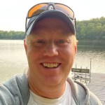 Selfie of Nick Lang smiling in a hat, gray hoodie, and white t-shirt. He's standing on a dock at a lake.