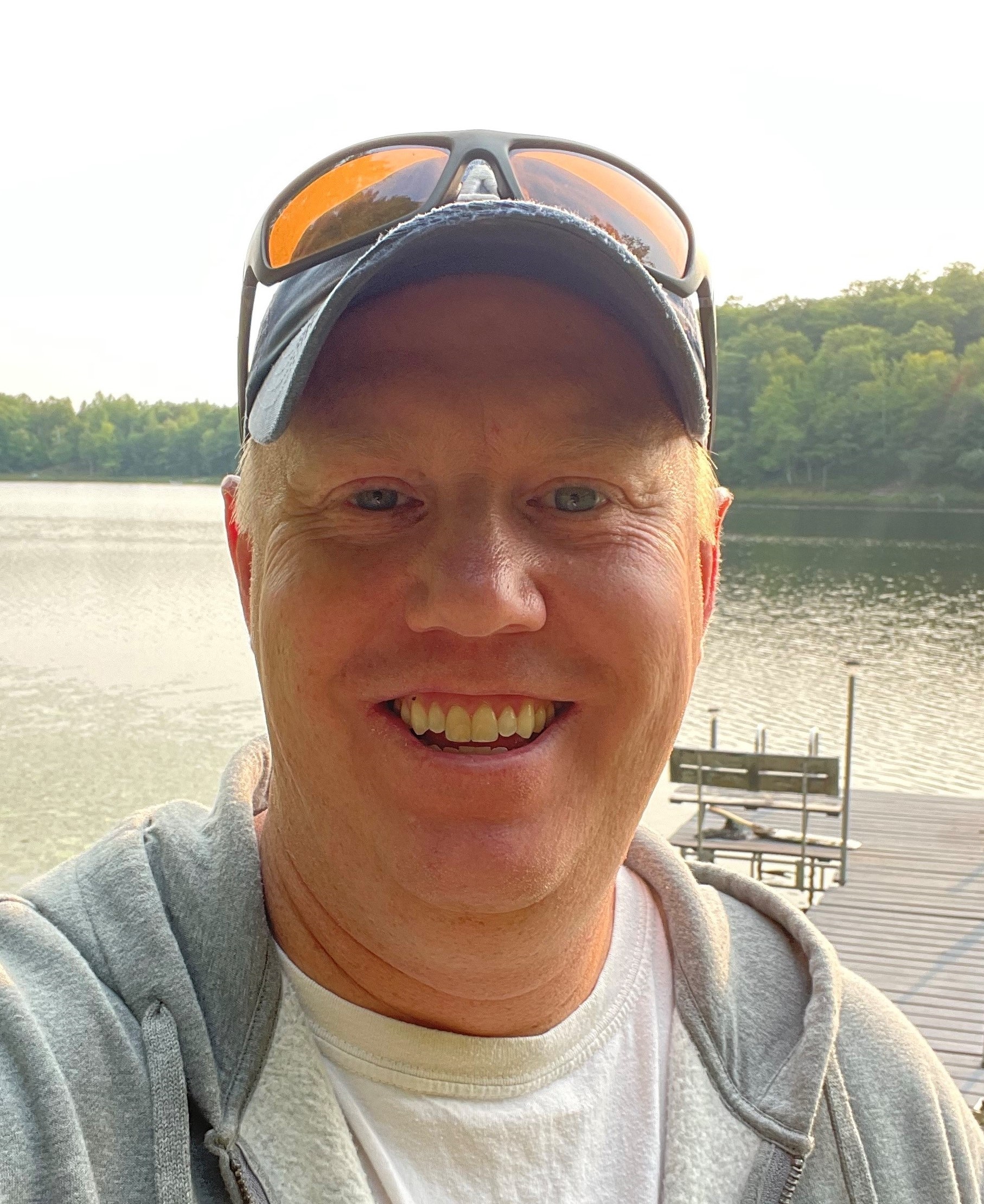 Selfie of Nick Lang smiling in a hat, gray hoodie, and white t-shirt. He's standing on a dock at a lake.