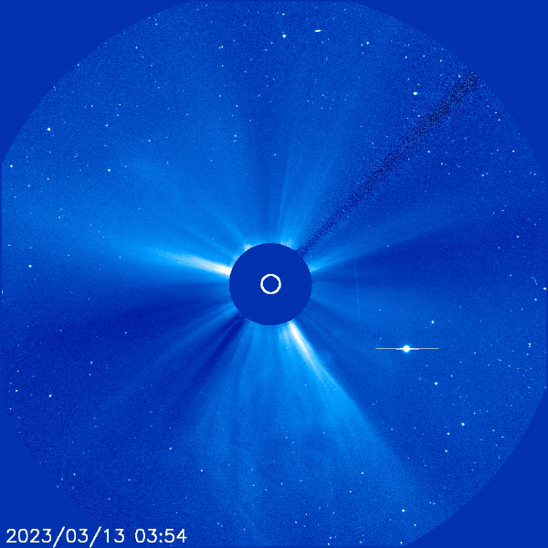 A blue image of the the Sun's atmosphere. In the middle is a dark blue circle, surrounding it are faint white streams of light. A large burst of light shoots out from the center in a circle into all directions.