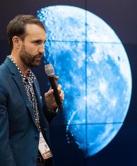bearded man with teal sport jacket holding microphone in front of hyperwall