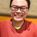 man with brushed back dark hair and glasses, broad smile, wearing orange v-necked sweather with thin black-with-white border stripe across chest, black piping at the neck, and yellow T-shirt underneath