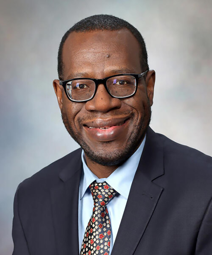 Portrait of an African American man with a short haircut, wearing dark-rimmed glasses. He is wearing a dark suit and multicolored tie.