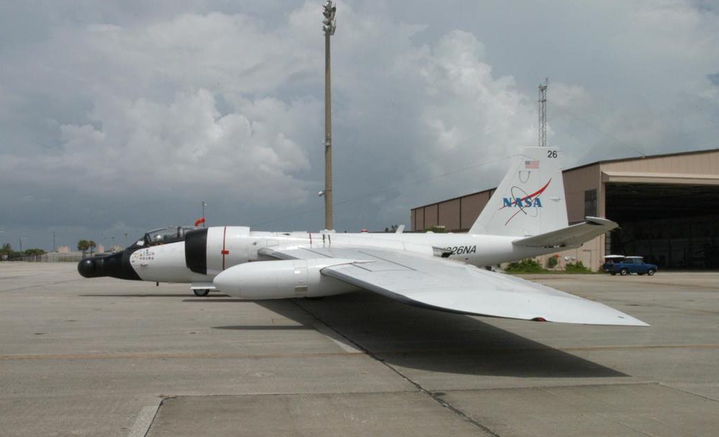 A small white airplane sis on a beige tarmac. It has a skinny nosecone and a fin that sticks straight up, with the NASA insignia.