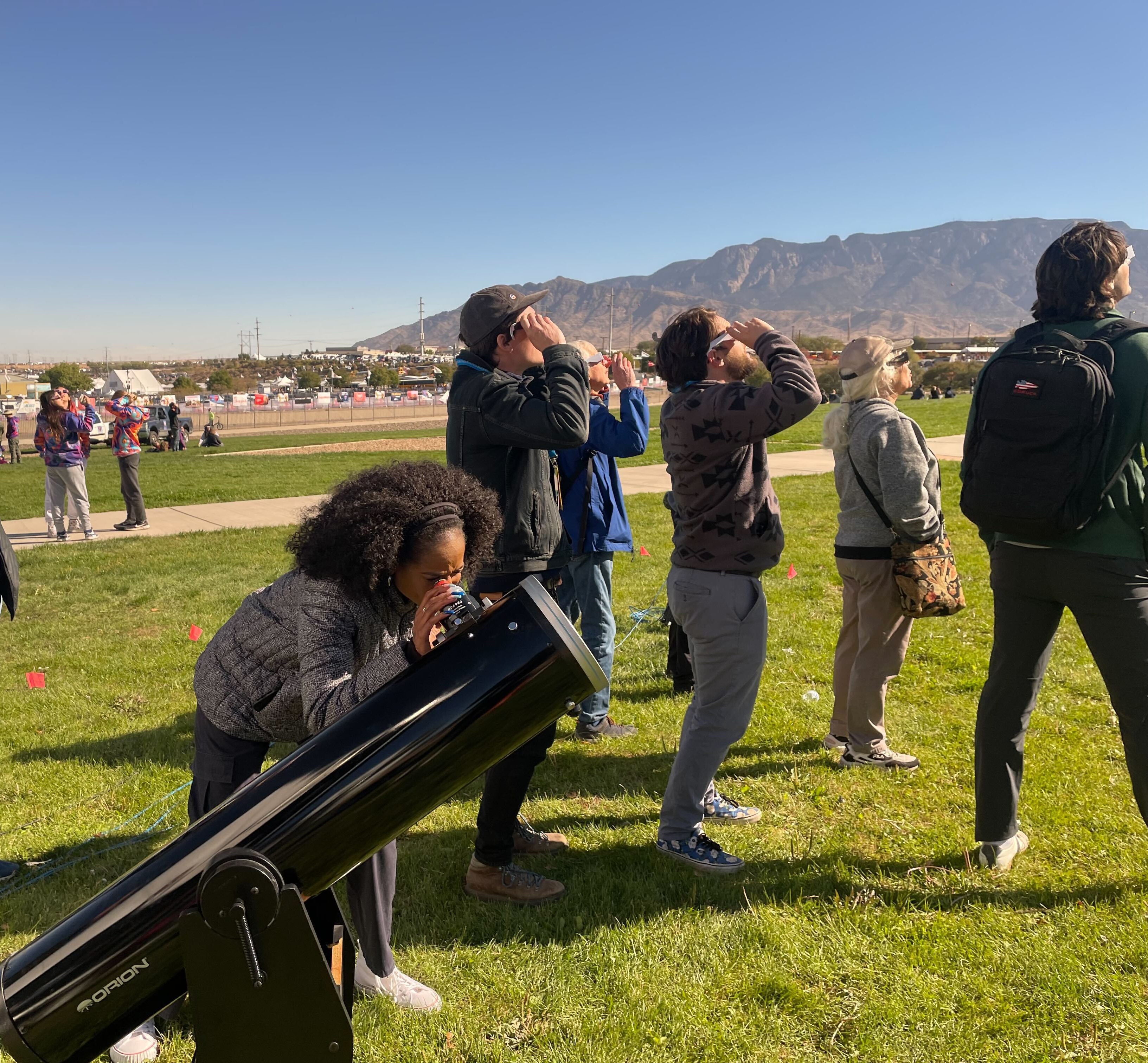 Against a landscape of a flat field and mountains, several people look toward the sky while wearing eclipse glasses. One woman looks through a telecope.