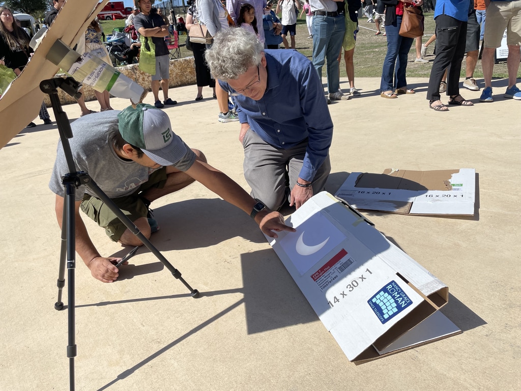 Two people kneel over a white piece of cardboard. Above it, a device is projecting an image of the crescent Sun onto cardboard.