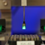 A thin robotic arm lifts a small glowing pellet from a tray of pellets.