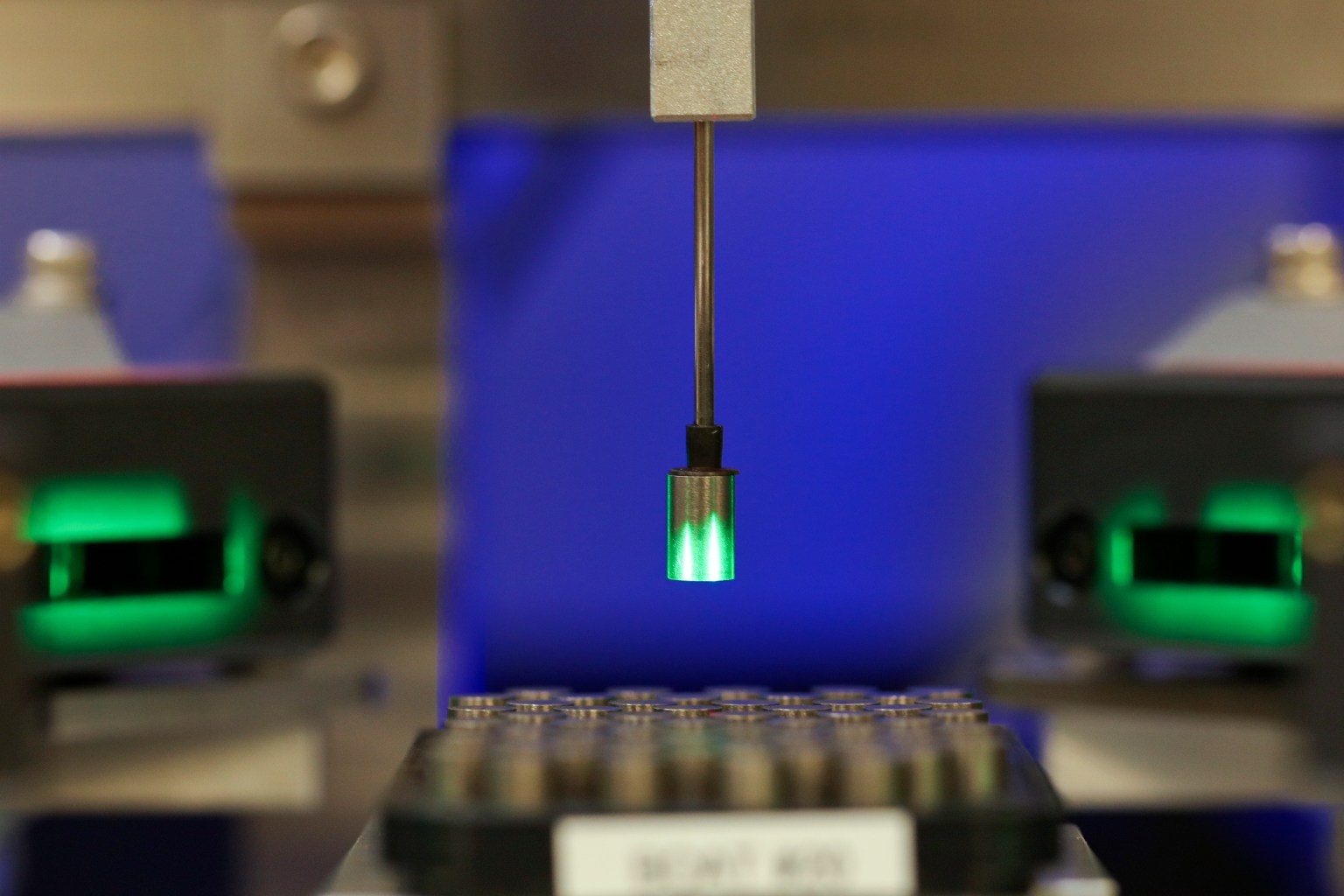 A thin robotic arm lifts a small glowing pellet from a tray of pellets.