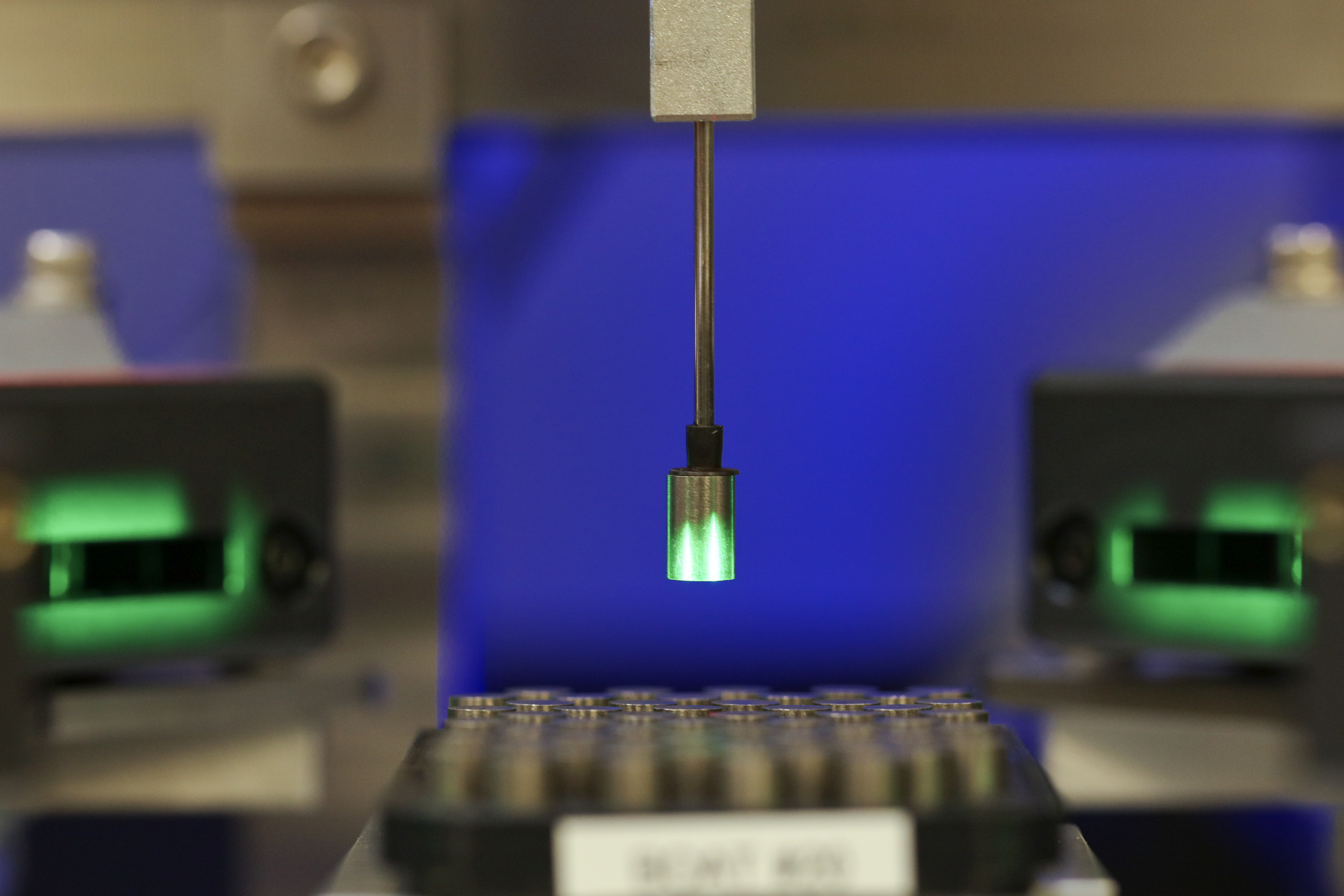 A thin robotic arm lifts a small glowing pellet from a tray of pellets.