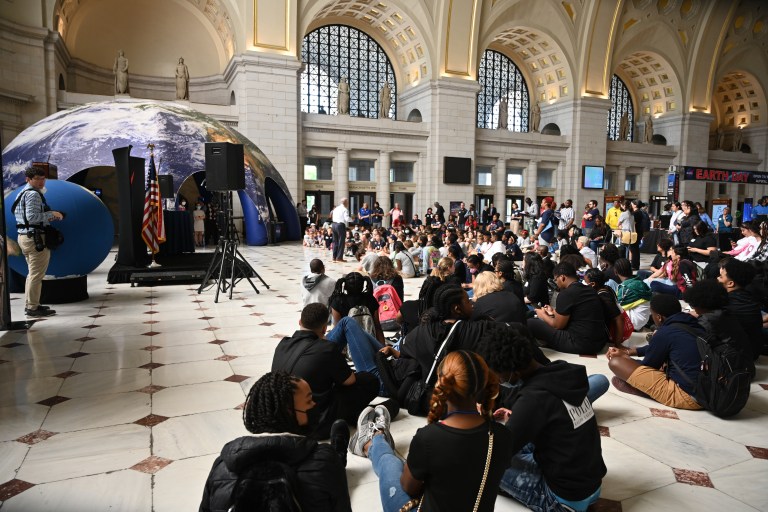 Numerous kids sit on tiled floor in a building with illuminated classical roman architecture style decor and high vaulted ceilings as they await a speaker to make their way to the temporary floor level staging and sound system. An inflatable Earth dome sits in the background with activities set up inside and an earth day banner hangs on the wall in the far right of the image