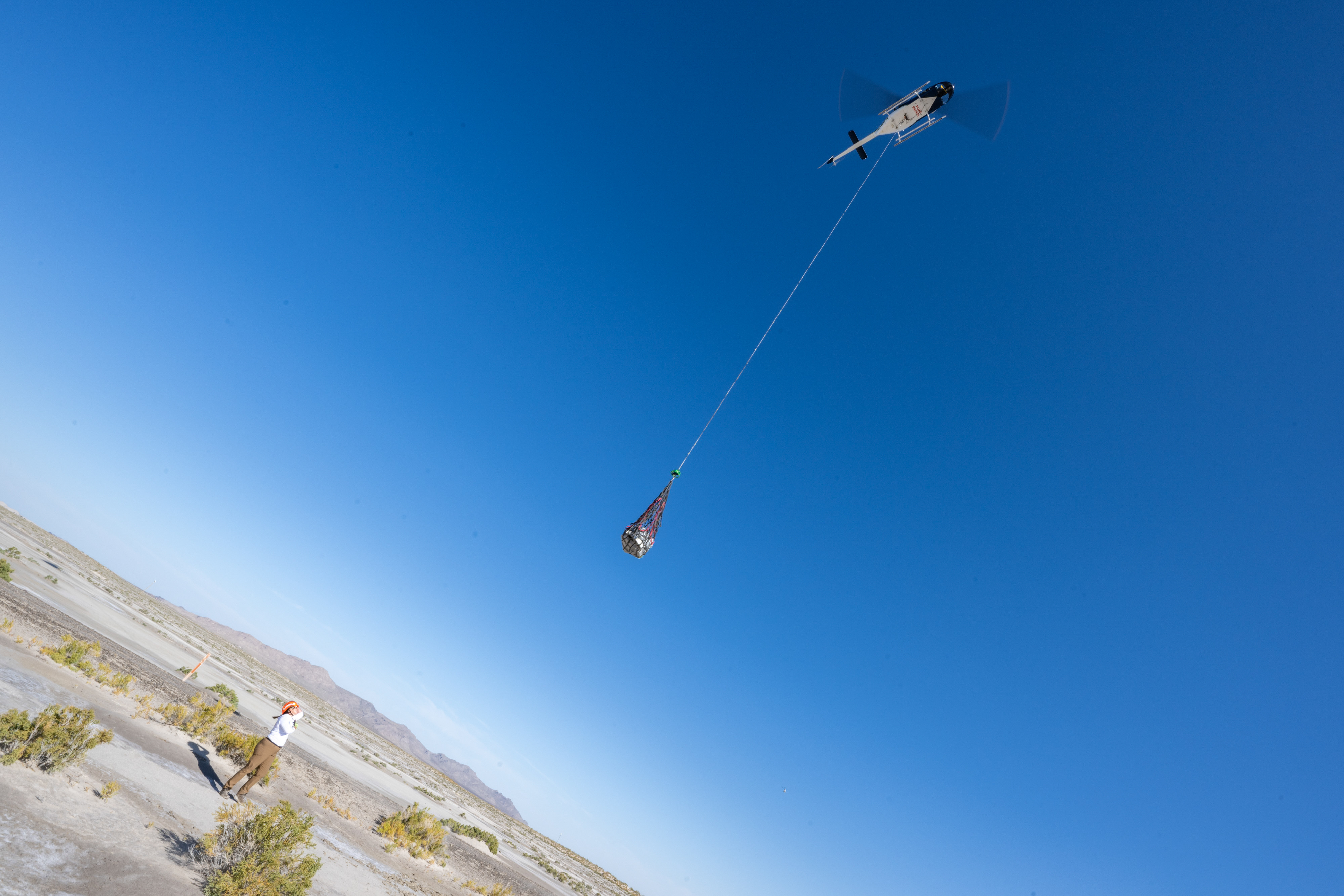 The sample capsule is suspended the helicopter by long line against a bright blue sky and the desert below. A woman looks up at the capsule as it departs.⁣