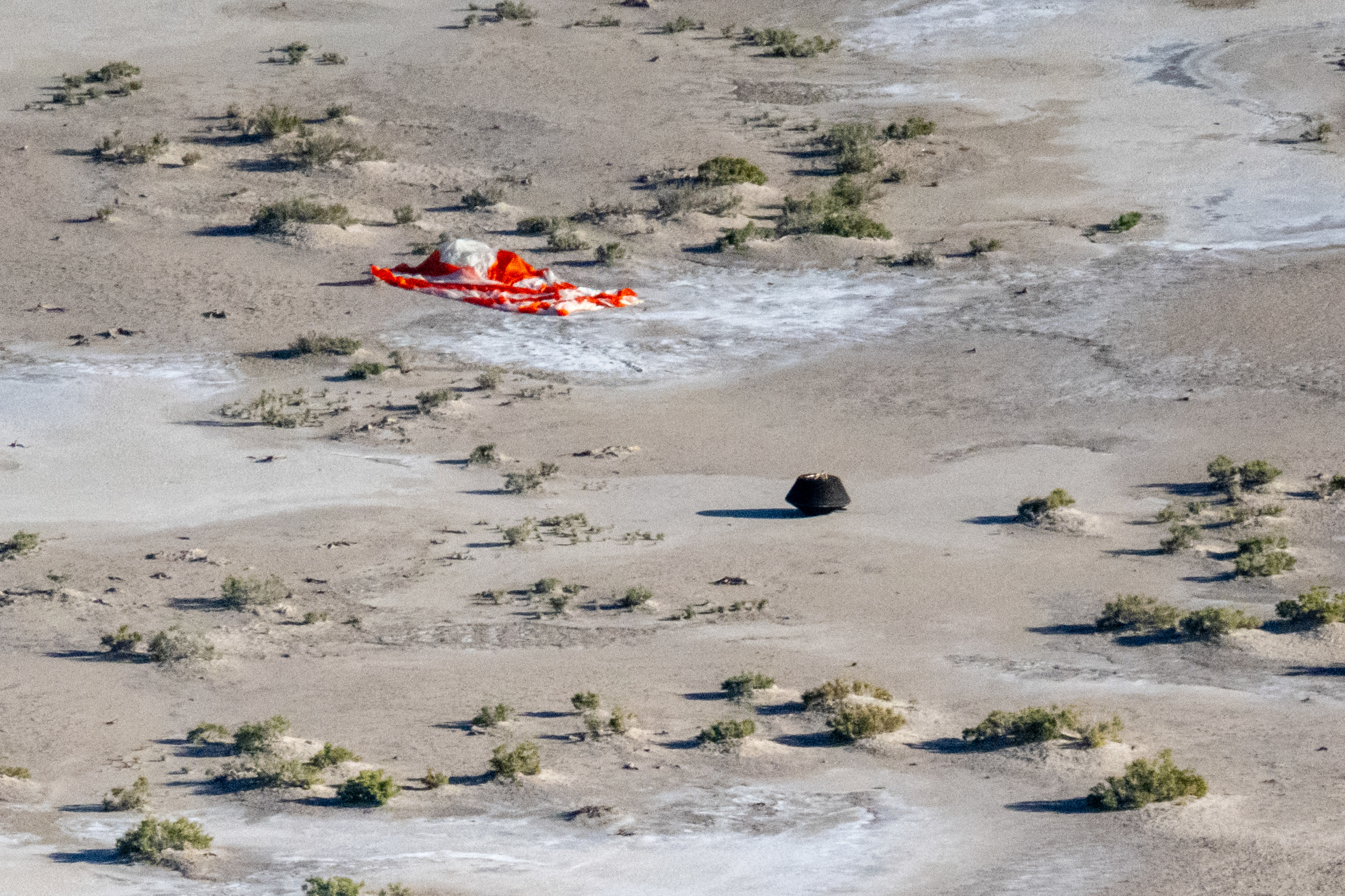 Aerial view of the sample return capsule on the desert ground with an orange-and-white parachute attached.⁣