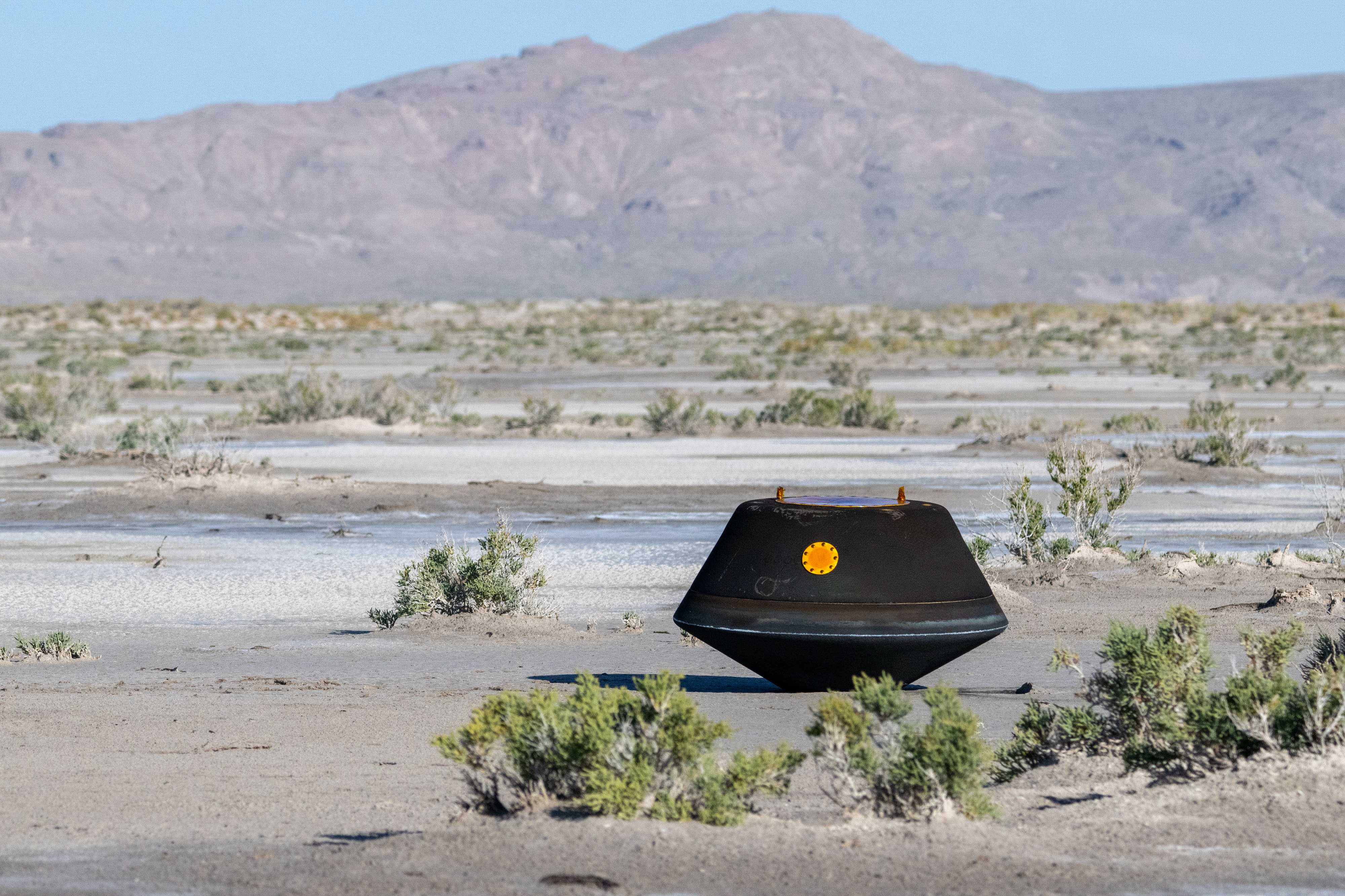 Black, top-shaped sample return capsule sits on the desert ground.⁣