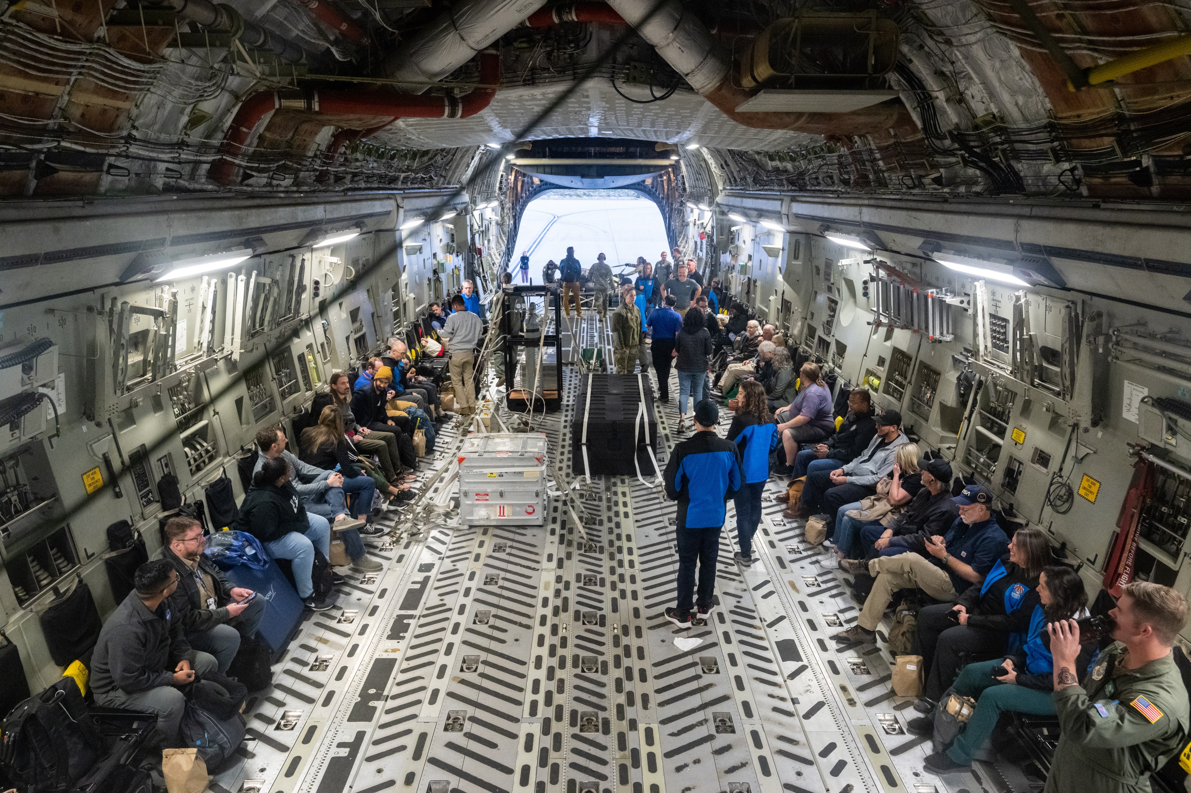 A group of people sit inside a C-17 Globemaster aircraft with the sample return capsule and its accompany nitrogen tanks in the middle of the plane