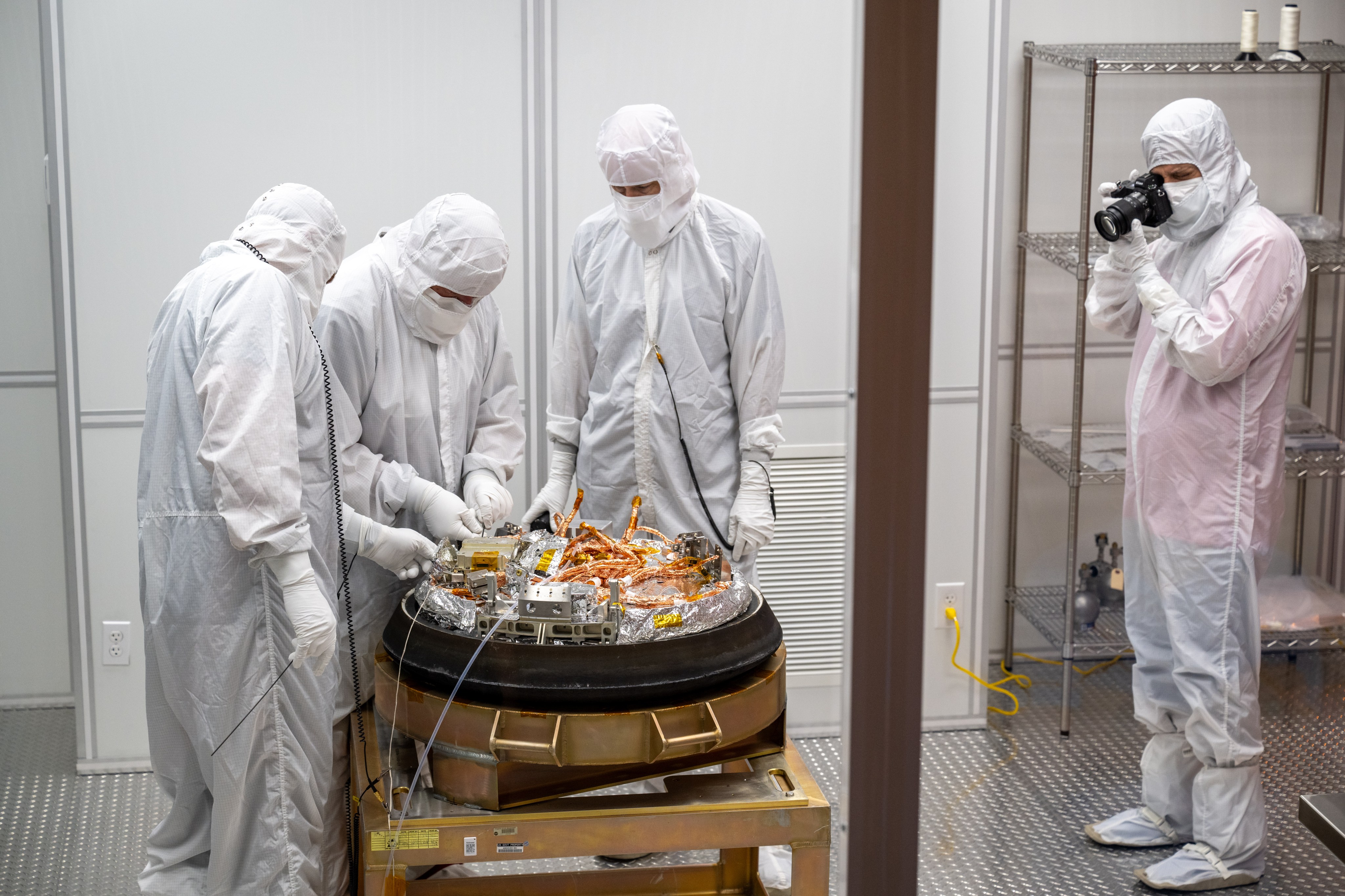 Three people in white cleanroom suits work on the capsule, one person in a white cleanroom suit takes photos