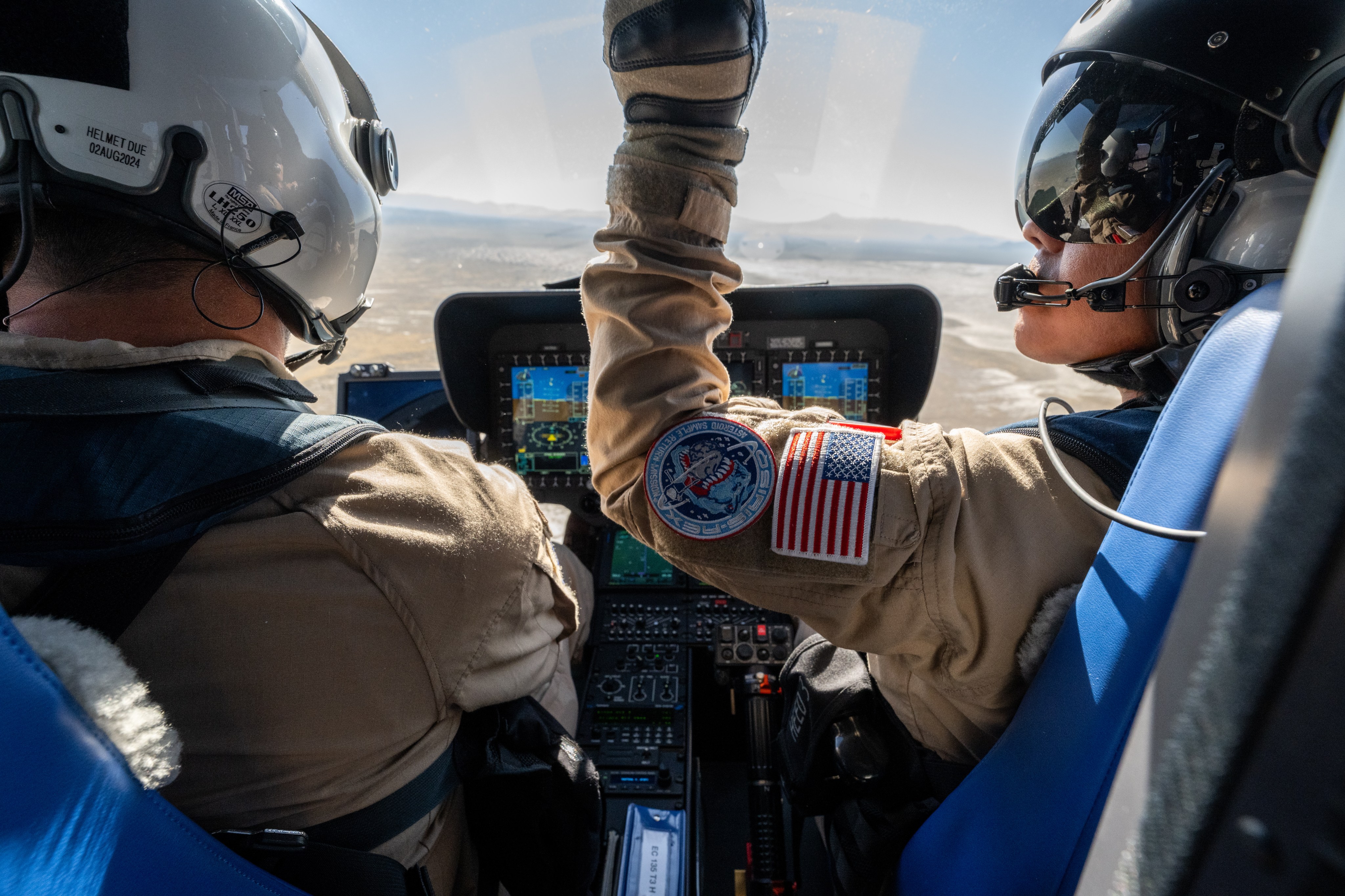 From behind, we see two people fly a helicopter. One lifts his arm, showing an American flag patch and OSIRIS-REx mission patch