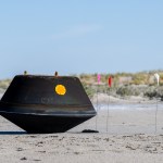 Black, top-shaped sample return capsule sits on the desert ground with flags surrounding it.