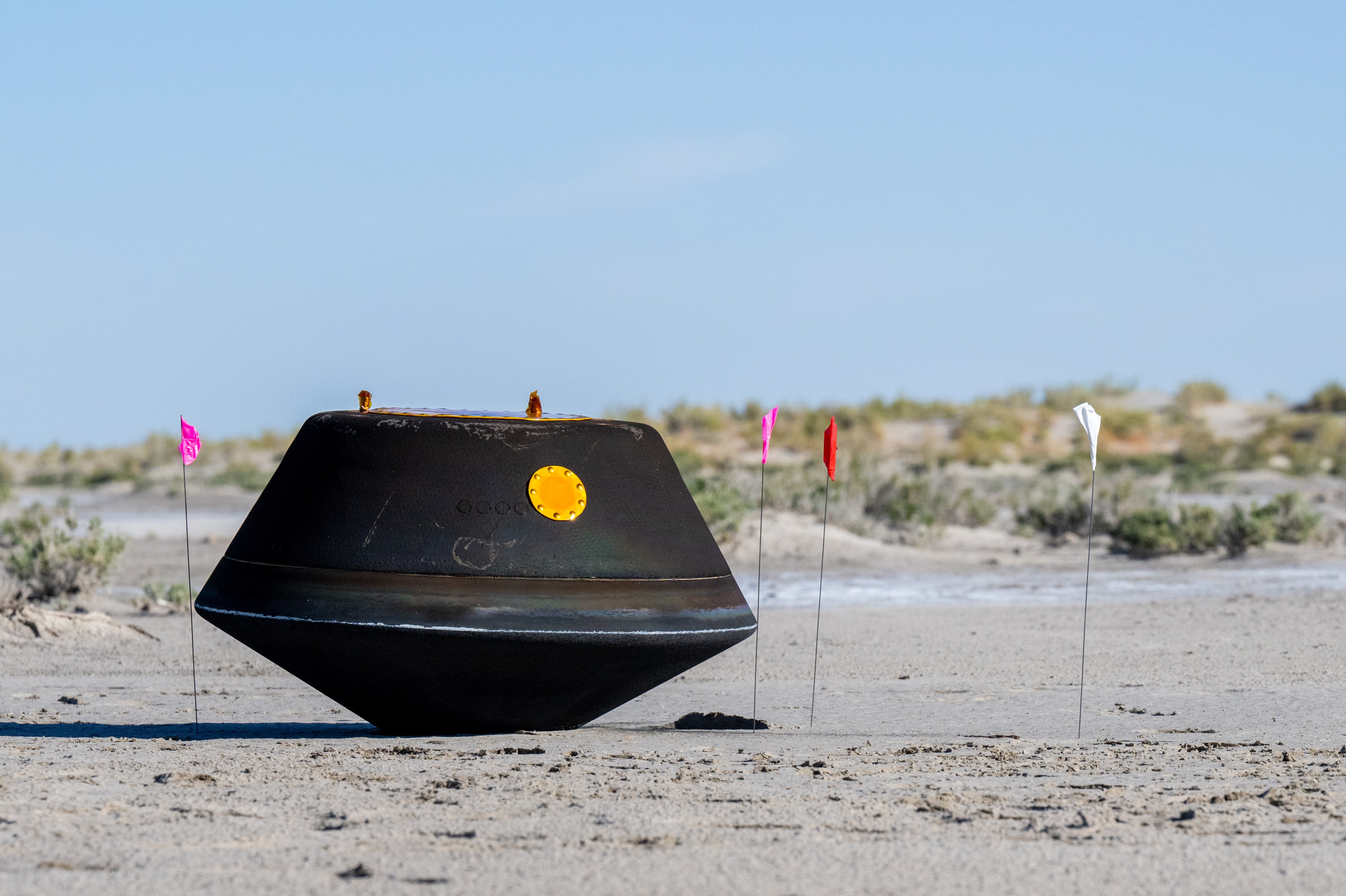 Black, top-shaped sample return capsule sits on the desert ground with flags surrounding it.
