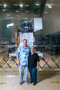 A man and a woman stand centered in the image, smiling at the camera. The man is taller than the woman and is wearing a blue button-up short sleeved polo with a Hawaiian shirt pattern of NASA logos, satellites, and clouds printed on it. The woman is wearing a black shirt and dark blue jeans. Behind them, positioned behind a protective sheet of plastic is the PACE spacecraft, which is covered in a silver foil like material.