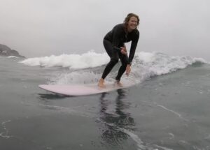 A woman stands on a pale pink surfboard, riding in a wave. She wears a black wet suit which covers her legs, torso, and arms. Her hair is wet. She is facing the right of the image, while traveling towards the left. The water she is surfing on is a dark gray color, with a white wave break behind her. The sky is cloudy and gray.
