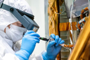 A woman stands in the left portion of the image. She is seen from the shoulders up and is wearing a white clean room suit that covers her torso, arms, and goes over her head. She also has on blue latex gloves, a white mask over her nose and mouth, and magnifying glasses. To the right of the image is a portion of the PACE spacecraft, which is covered in a gold-colored foil-like material. The woman is holding wiring from the spacecraft and is looking closely at it.