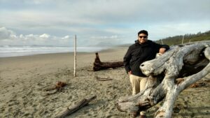 A man wearing khaki pants, a black sweatshirt, and sunglasses stands to the right of the image. He is leaning against a large piece of driftwood and standing on the beach. There are three other smaller pieces of driftwood scattered throughout the picture. Majority of the picture is taken up by the beach, but the top of the image shows the horizon, some clouds, and a blue-gray colored sky.