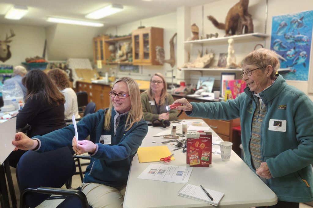 Three women are in focus in the front of the image. They are gathered at a white six-foot table in a small room crowded with other people. The shelves around the small room are full of science materials and equipment. All of the people in the image are busy creating eclipse activities with craft materials. The woman in focus on the left is seated and holding a pinhole eclipse viewer that she has just made from materials scattered across the table. The woman in focus on the right is standing and smiling, admiring the pinhole viewer. The woman in focus in the center of the image is seated and is also smiling and admiring the pinhole viewer.