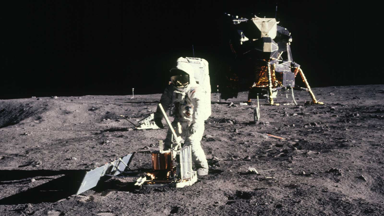 An astronaut in a spacesuit installs an experiment on the surface of the Moon. The lunar landing module is visible in the background.