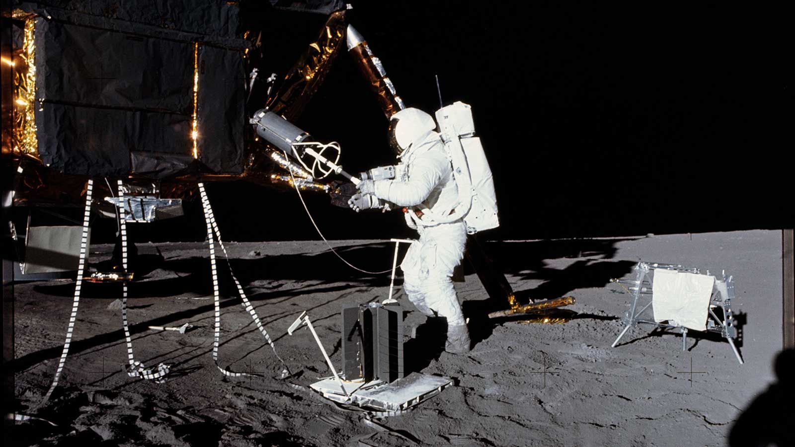 An astronaut in a spacesuit removes a cylinder from the side of a lunar lander on the Moon.