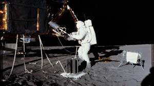 An astronaut in a spacesuit removes a cylinder from the side of a lunar lander on the Moon.