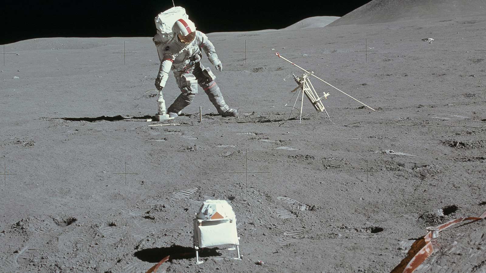 An astronaut in a spacesuit leans over to install an experiment on the Moon.