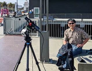 Photo of a man sitting on a bench outside next to a camera on a tripod