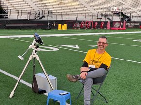 Photo of a man in a yellow tshirt sitting in a chair on a football field next to a tripod and camera.