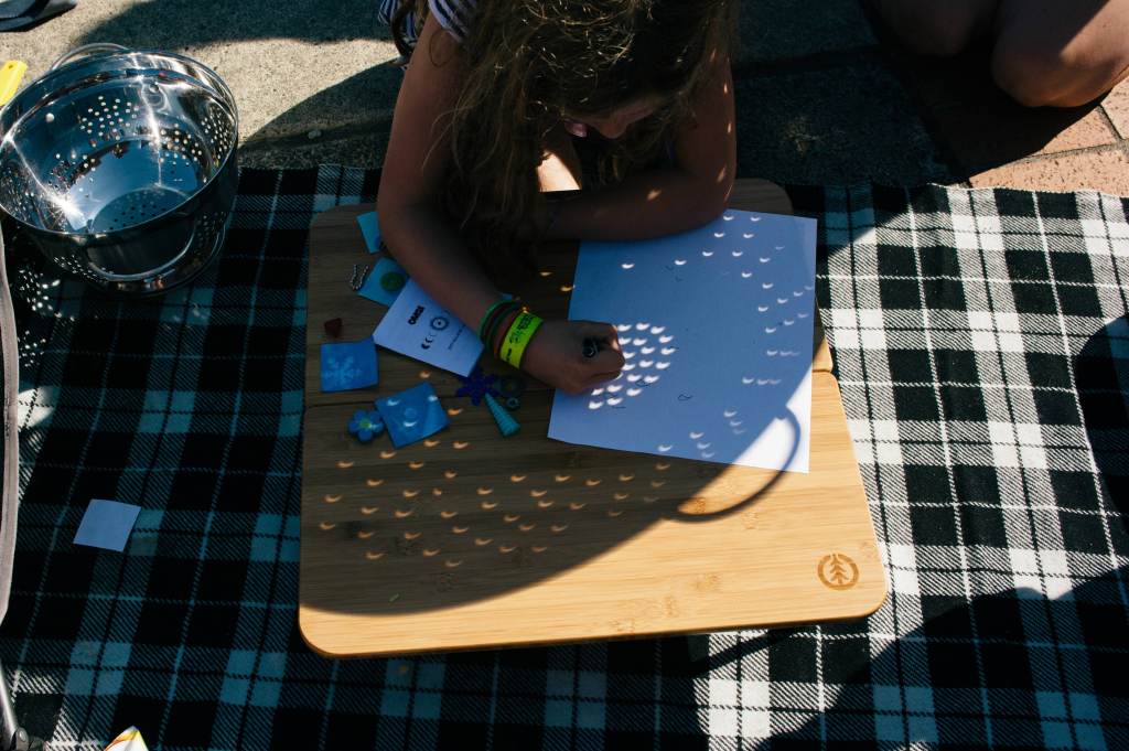 Photo of a person tracing the crescent-shaped light shining onto a piece of paper through a colander during a partial solar eclipse.