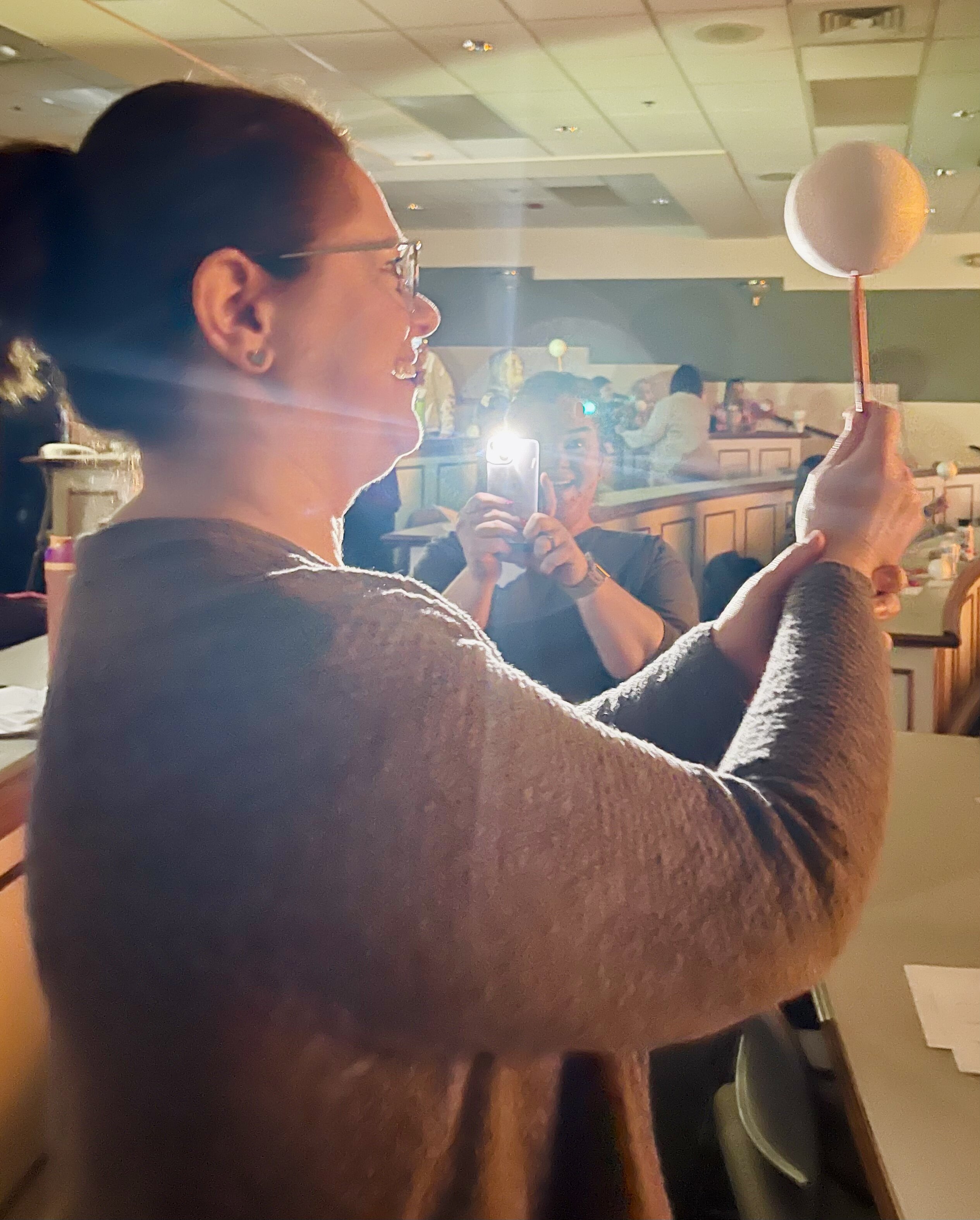 Photo of two teachers as they model the sun/moon/earth relationship with flashlights and styrofoam ball. One teacher sees the moon phases and smiles.