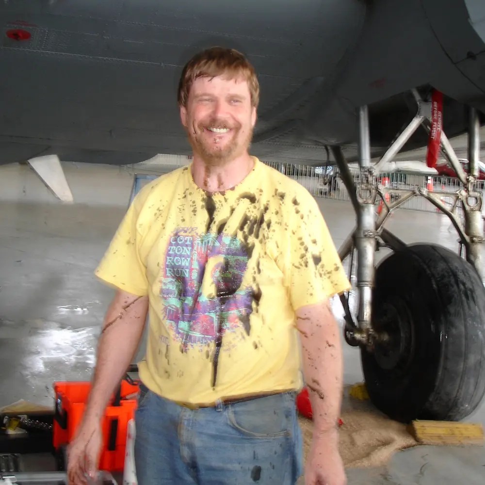 Photo of a smiling man wearing a yellow tshirt that is splattered in mud.