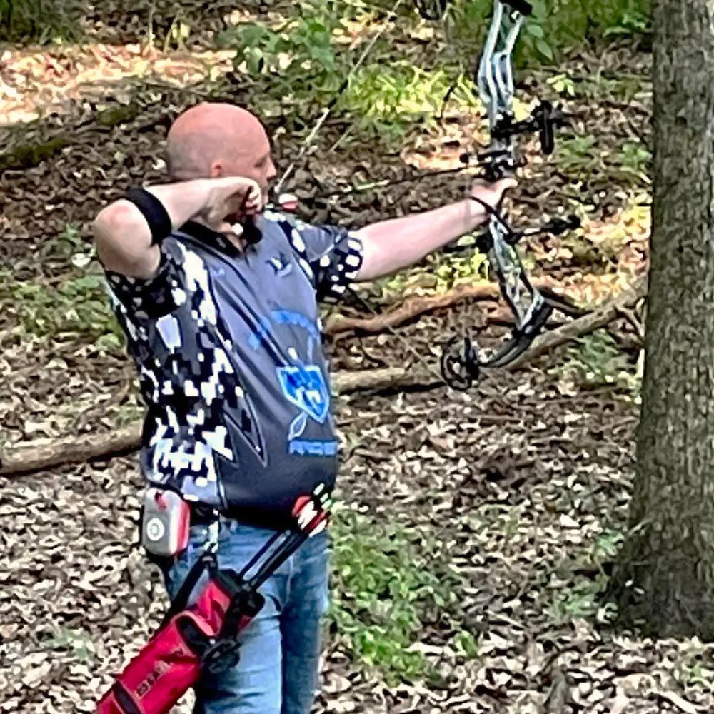 Photo of a man in the woods shooting a bow and arrow.