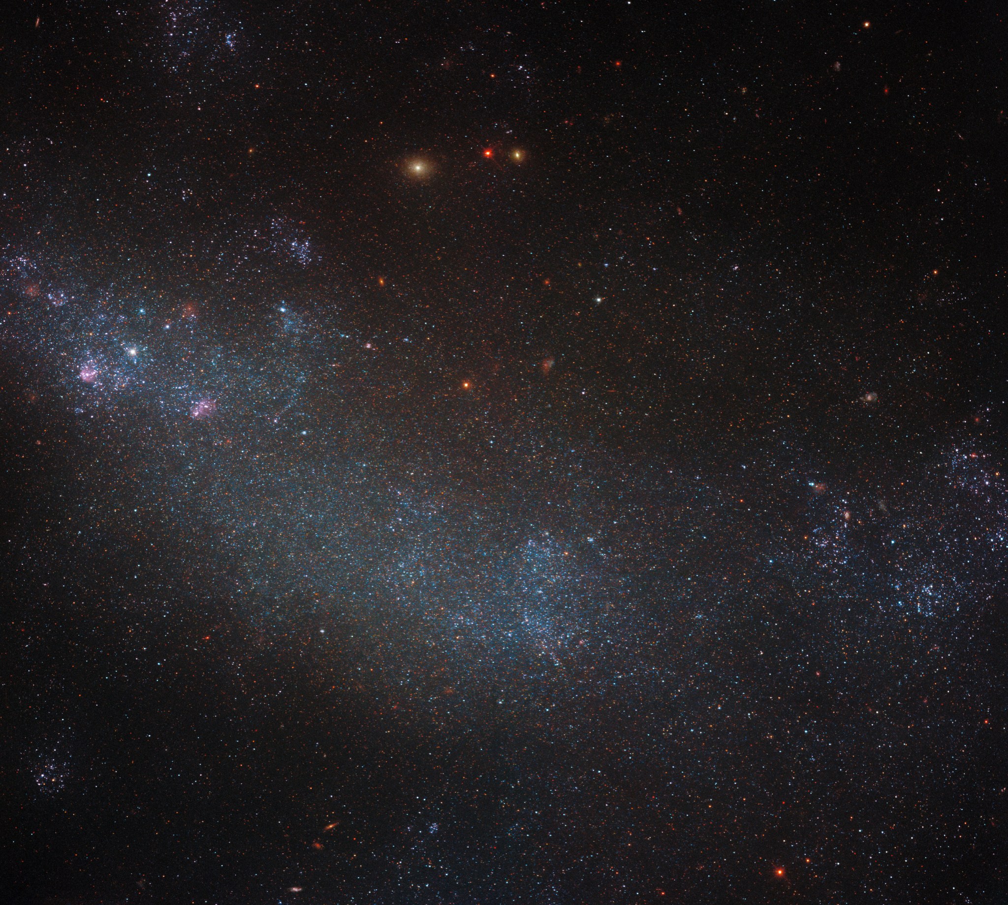 An irregular galaxy: a cloud of tiny, point-like stars on a dark background. The cloud is densest along a broad, curved band across the center of the image. It is colored a faint blue with glowing purplish patches, and the stars grow less dense out to the edges but don't fully vanish. A few distant background galaxies appear among the stars as glowing spots.
