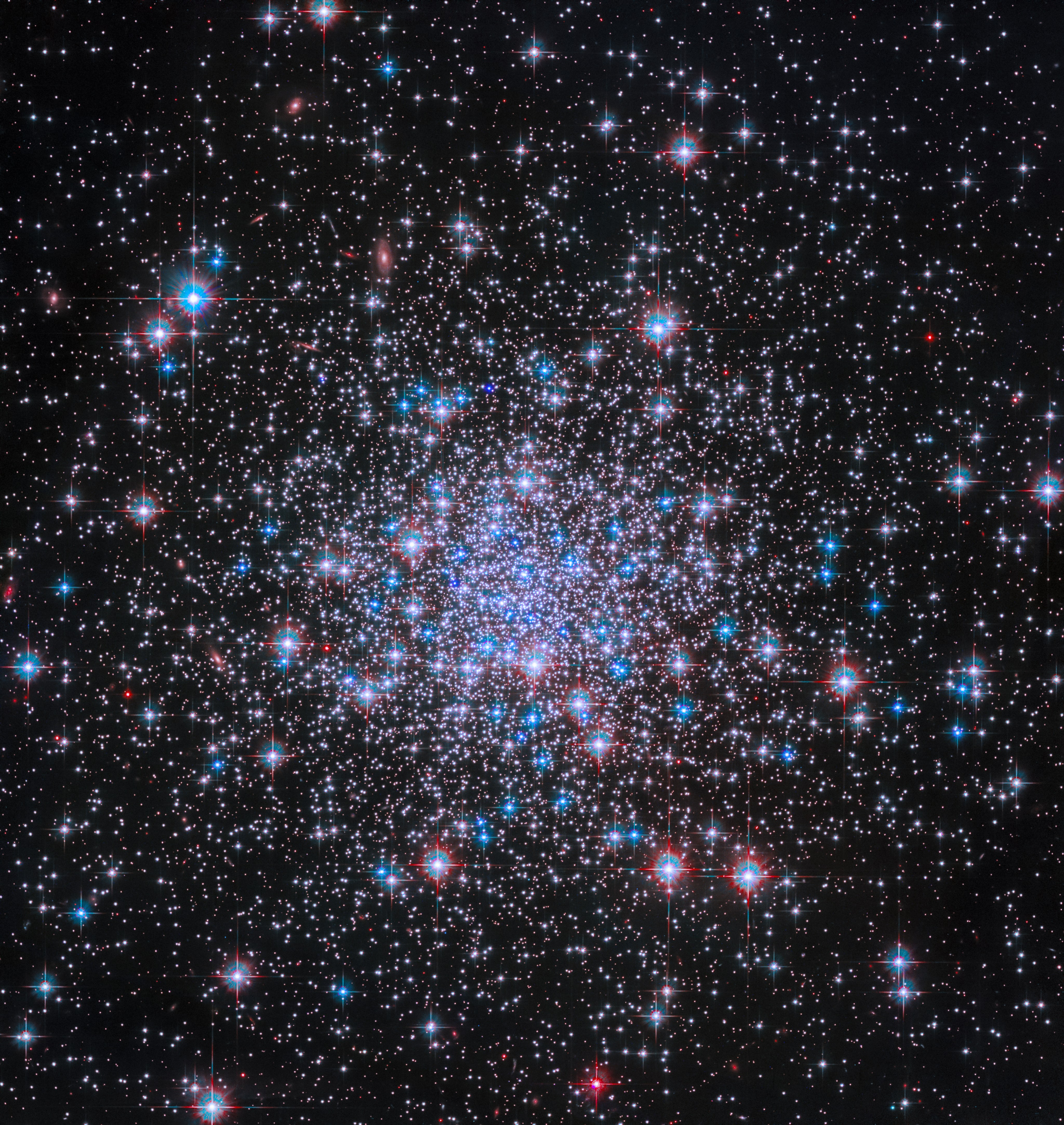 Thousands of bright stars shine against black space, more tightly condensed near the image’s center. The stars glint in shades of white, blue, and red, and diffraction spikes are visible around the foreground stars.